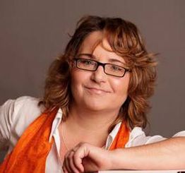Eine lächelnde Person mit Brille und orangenem Schal vor grauem Hintergrund.