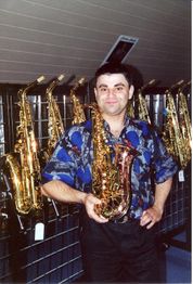 Mann in gemustertem Hemd hält ein Saxophon vor einem Regal mit mehreren Saxophonen.