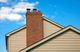 Brick chimney on a beige house roof against a clear blue sky with a few clouds.