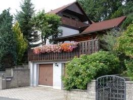 Haus mit Balkon mit bunten Blumen, Garage und Garten, umgeben von Bäumen und Sträuchern.