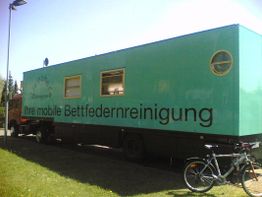 Grüner LKW-Anhänger mit Aufschrift „Ihre mobile Bettfedernreinigung“, daneben steht ein Fahrrad.