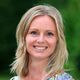 Vrouw met blond haar en kleurrijke blouse lacht naar de camera in groene omgeving.
