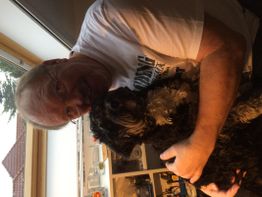 Ein Mann in einem weißen T-Shirt umarmt einen kleinen schwarzen Hund in einem Raum mit Fenster.