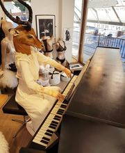 Person mit Hirschkopf spielt Klavier in Schaufenster, umgeben von Schaufensterpuppen mit Unterwäsche.