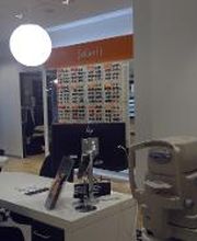 Optiekwinkelinterieur met tafels, stoelen en meetapparatuur; brilposters op de achtergrond.