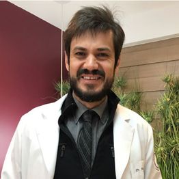 Homem sorridente de jaleco branco, gravata escura. Ao fundo, parede vermelha e plantas.