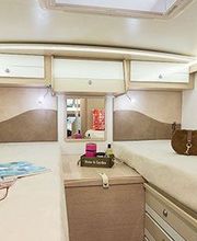 Interior de una caravana con dos camas, revista, gafas, bolsa, y decoración sencilla.