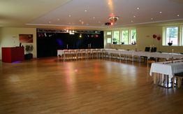 Großer Raum mit Holzfußboden, gedeckte Tafel, Bühne und Dekorationen für eine Veranstaltung.