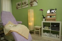 Wellnessraum mit Liege, Regal mit Handtüchern, Blumen und Lampe vor grüner Wand.