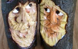 Zwei geschnitzte Gesichter in Holzscheiten, nebeneinander mit weißen und orangefarbenen Details.