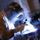Welder working with bright sparks, wearing protective gear and mask in a dimly lit workshop.