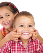 Niño y niña sonrientes, ella estira las mejillas del niño. Fondo blanco.