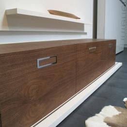 Holzkommode mit schlichten Griffen, darüber ein Regalbrett auf weißer Wand, moderner Stil.