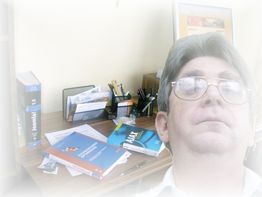Ein Mann mit Brille, im Hintergrund ein unaufgeräumter Schreibtisch mit Büchern und Schreibutensilien.