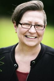 Eine lächelnde Person mit Brille und kurzem Haar vor grünem, unscharfem Hintergrund.