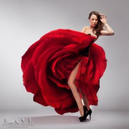 Frau in rotem Kleid, das wie eine Blume aussieht, posiert anmutig vor grauem Hintergrund.