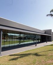 Prédio moderno com fachada de vidro e jardim com palmeiras ao lado, em dia ensolarado.