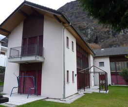 Modernes zweistöckiges Haus mit braunen Fensterläden vor einem felsigen Hügel und Rasenfläche.