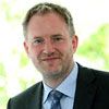 Mann mit Anzug und Krawatte lächelt und steht vor einem unscharfen grünen Hintergrund.