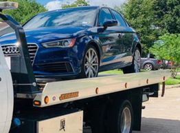 Blue car on a tow truck, parked outdoors under a clear sky with trees in the background.