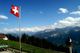 Schweizer Flagge weht vor Alpenpanorama mit Berggipfeln und blauem Himmel im Hintergrund.