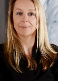 Eine Frau mit blonden Haaren lächelt leicht in die Kamera, sie trägt schwarze Kleidung.