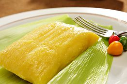 Pamonha em folha de milho com garfo e pimenta colorida ao lado.