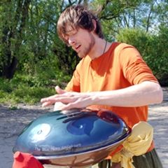 Mann spielt im Freien ein Handpan-Instrument, trägt ein orangefarbenes Hemd.