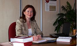 Femme assise à un bureau, écrivant, avec ordinateur portable et livres autour d'elle.