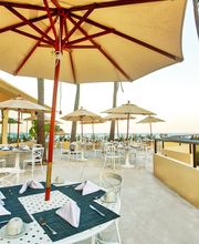 Terraza con mesas bajo sombrillas, vista al mar y palmas. Ambiente relajado y soleado.