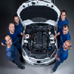 Six mechanics in blue uniforms stand around an open car hood, smiling at the camera.