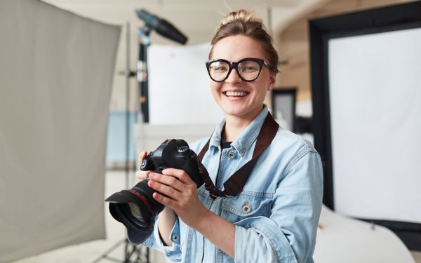 Eine junge Fotografin in einem Jeanshemd und mit einer Brille, hält lächelnd eine Kamera in der Hand, um die Bilder zu kontrollieren, die sie eben gemacht hat.