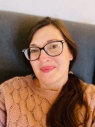 Person mit Brille lächelt in die Kamera, trägt einen rosa Pullover und sitzt vor grauem Hintergrund.