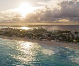 Vista aérea de una playa con hoteles al atardecer, cielo nublado y océano en calma.