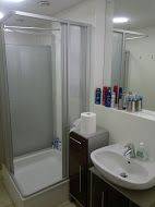 Kleines Badezimmer mit Dusche, Waschbecken, Spiegel und Kosmetikartikeln auf einem Schrank.