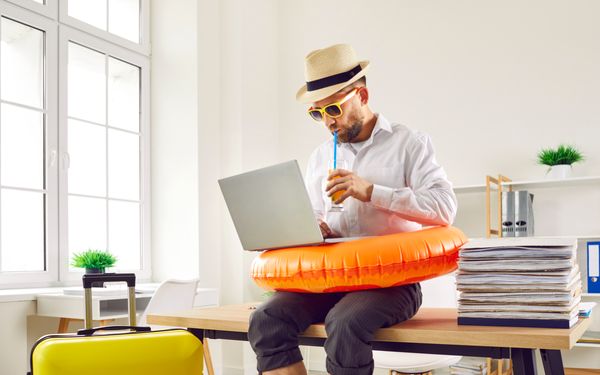 Mann arbeitet nach dem Urlaub mit Laptop, Sonnenhut, Getränk und Schwimmring im Büro.