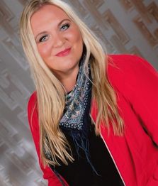 Eine lächelnde Frau mit blonder Haaren, rotem Blazer und Schal vor gemustertem Hintergrund.