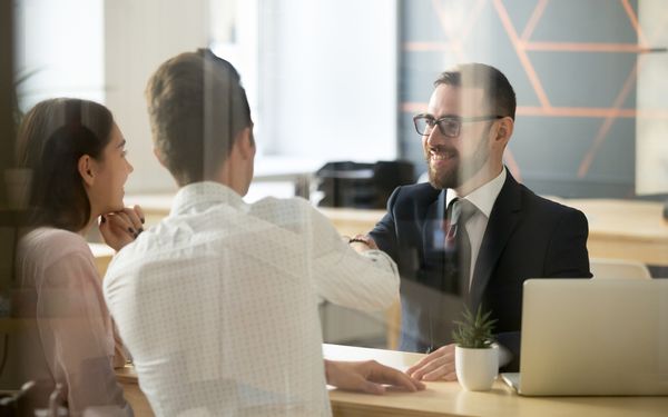 Ein Bankberater in einem dunklen Anzug und ein Mann in einem weißen Hemd, schütteln sich die Hände. Neben dem Mann im weißen Hemd sitze eine Frau mit langen glatten dunklen Haaren.