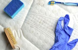 Cleaning tools on a mattress: sponge, brush, toothbrush, and blue gloves.