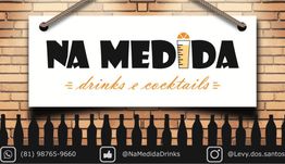 Placa "Na Medida" com contatos de mídia social e telefone, sobre fundo de tijolos com garrafas.