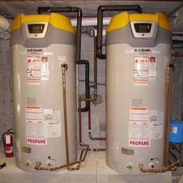 Two propane water heaters with digital displays, pipes, and safety labels in a utility room.