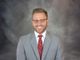 Smiling man in glasses and gray suit with red tie against a gray background.