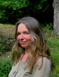 Eine Frau mit langen Haaren steht draußen vor einem grünen, bewaldeten Hintergrund.