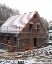 Einfamilienhaus im Bau mit Gerüst, umgeben von Schneelandschaft und Bäumen im Hintergrund.