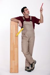 Ein Mann in Arbeitskleidung lehnt an einem Holzbrett und hält einen roten Stift nach oben.