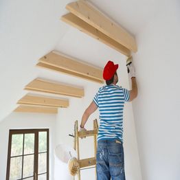 Person on ladder painting ceiling beams in a bright room with a window.