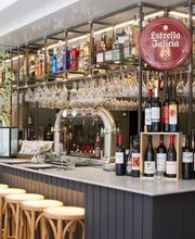 Bar moderno con estantes llenos de botellas y taburetes de madera. Letrero de Estrella Galicia.