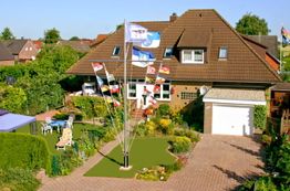 Backsteinhaus mit Garten, Flaggenmast und Garage in einer grünen Wohngegend bei sonnigem Wetter.