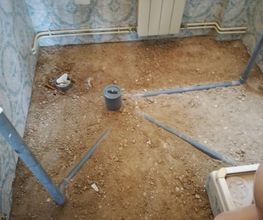 Pisos de tierra y tuberías grises en una habitación sin terminar, con radiador al fondo.