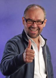 Mann mit Brille und Bart zeigt Daumen hoch, lächelt vor violettem Hintergrund.